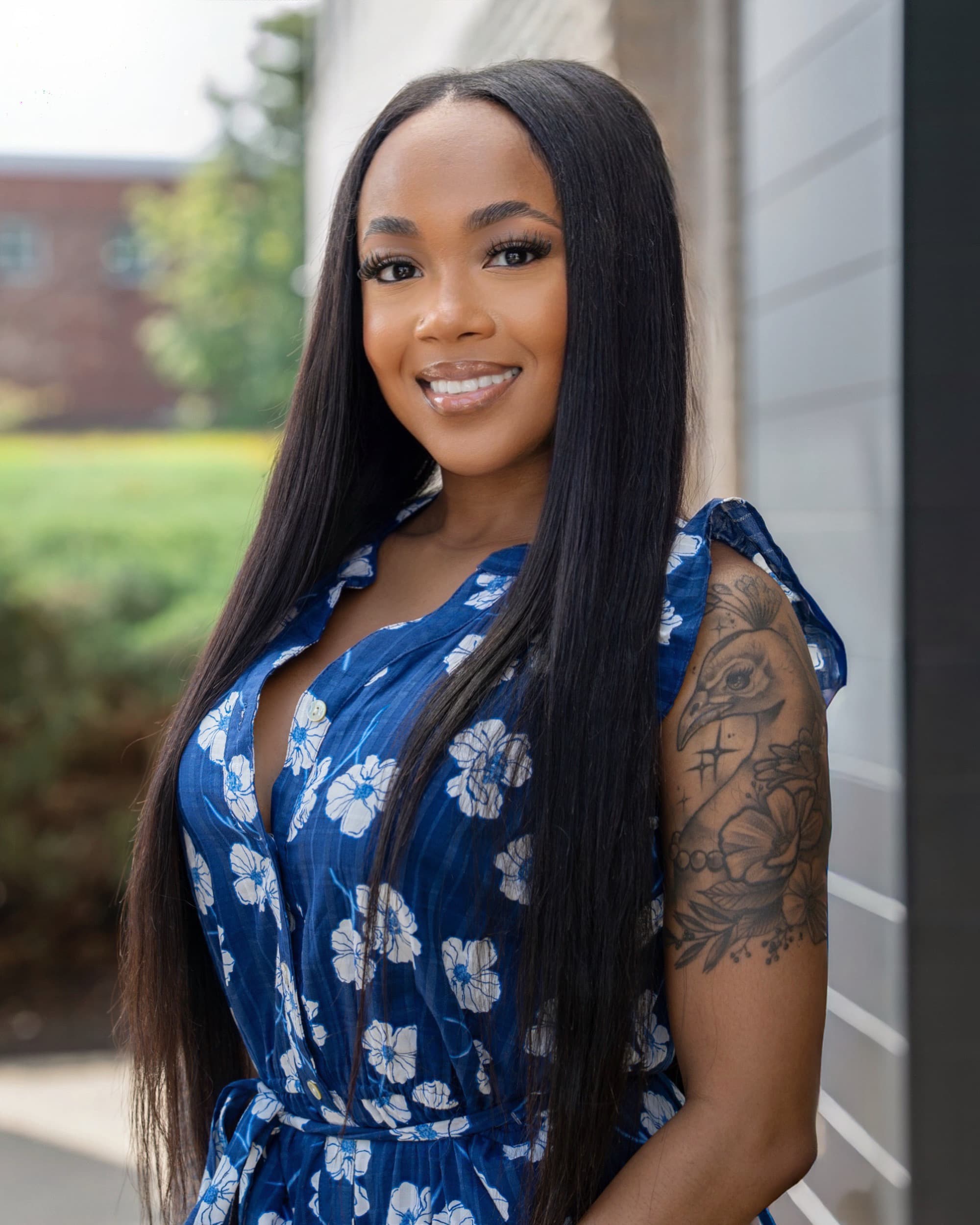 Smiling Black woman with long straight hair and arm tattoo in a blue floral dress.